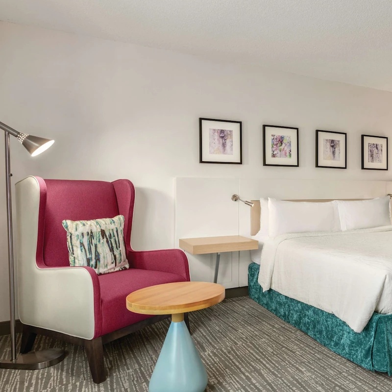Brand-compliant HGI wingback lounge chair in red and cream upholstery, paired with a Studio Teak activity table for a modern Hilton Garden Inn guestroom interior.